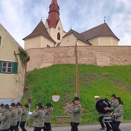 Eine Marschkapelle in Uniformen mit Hüten und Stiefeln tritt vor einer Kirche mit einem Turm auf der Straße auf.