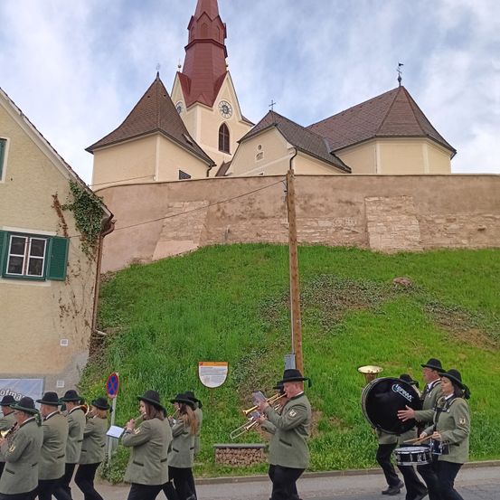 Eine Marschkapelle in Uniformen mit Hüten und Stiefeln tritt vor einer Kirche mit einem Turm auf der Straße auf.