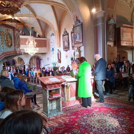 Ein Kircheninneres mit einer grün gekleideten Frau, die am Altar steht, einem Mann neben ihr und einer Menge von Menschen, die auf den Kirchenbänken sitzen.