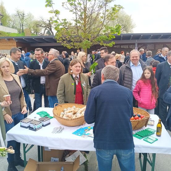 Eine Gruppe von Menschen steht um einen Tisch mit Essen in Körben, darunter ein älterer Mann, der mit einer Frau und einem Kind spricht, während andere unter sich reden.