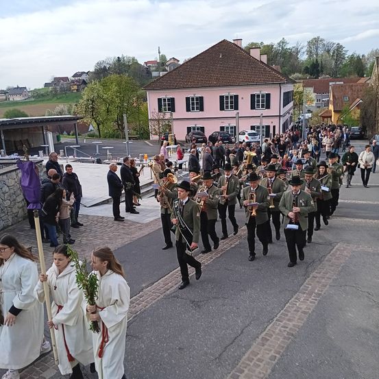Eine Gruppe von Menschen in grünen Uniformen und Hüten marschiert die Straße entlang und hält Musikinstrumente. Einige Leute schauen von der Seite zu, und ein Gebäude mit vielen Fenstern ist im Hintergrund.