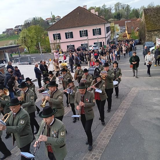 Eine Brassband aus Männern und Frauen in Uniformen marschiert durch eine Dorfstraße, begleitet von Zuschauern.