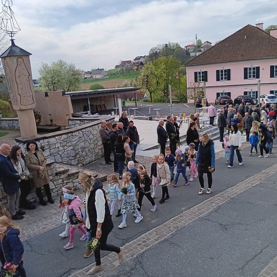 Eine Menschenmenge geht auf einem gepflasterten Weg, wahrscheinlich auf dem Weg zu einer Kirche. Sie tragen Blumen und Winterkleidung. Links steht eine Statue der Jungfrau Maria auf einem Betonpfeiler. Dahinter befindet sich ein Haus mit rosa Außenfassade und vielen Fenstern.
