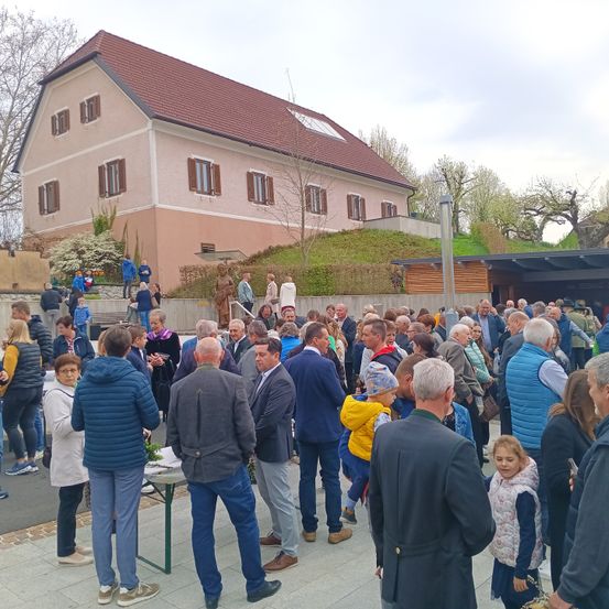 Eine große Gruppe von Menschen versammelt sich vor einem Haus. Sie sind in Winterkleidung gekleidet und einige von ihnen tragen Taschen. Im Vordergrund ist ein Tisch aufgebaut, auf dem Pflanzen stehen.
