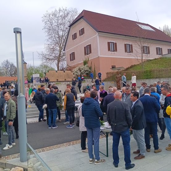 Eine große Menschenmenge versammelt sich draußen, einige stehen, andere sitzen. In der Mitte der Gruppe steht ein Tisch mit Pflanzen. Dahinter befindet sich ein Gebäude mit einem roten Dach und vielen Fenstern.