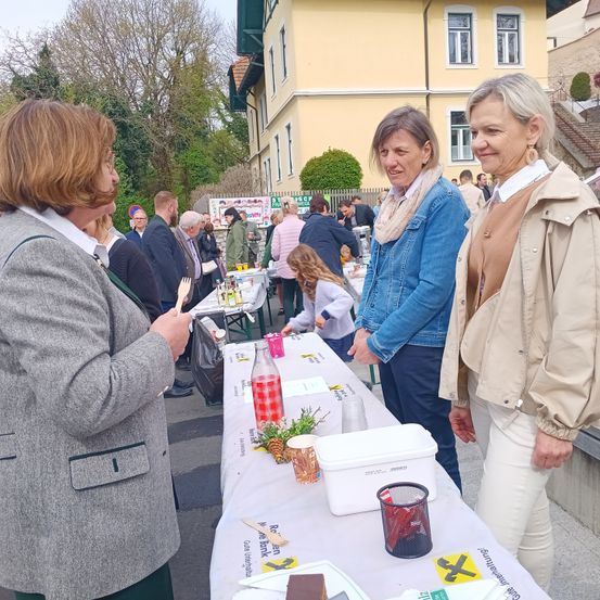 Eine Gruppe von Frauen steht vor einem Tisch mit Essen und Getränken, eine Frau hält eine Plastikgabel. Im Hintergrund stehen mehrere Personen vor Tischen. Die Gebäude im Hintergrund haben eine gelbe Fassade.