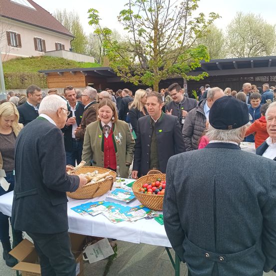 Eine große Gruppe von Menschen steht um einen Tisch herum und diskutiert miteinander. Einige tragen Körbe mit Essen und andere haben Tassen in den Händen. Dahinter befinden sich ein Haus, ein Schuppen und ein Baum.
