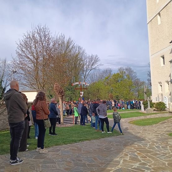 Eine große Menschenmenge versammelt sich während einer Tagesveranstaltung vor einer Kirche. Sie stehen auf einem Steinweg, in der Nähe eines Rasenbereichs, mit einem Kreuz im Hintergrund. Einige Menschen tragen Mützen und Turnschuhe.