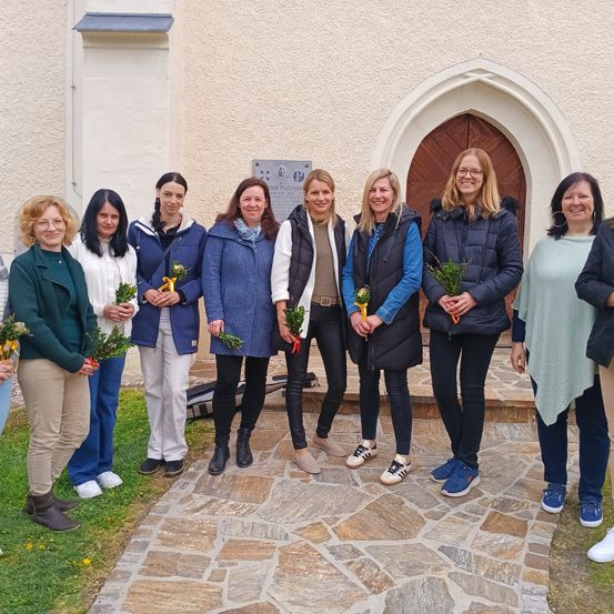 Acht Frauen stehen vor einem Gebäude und lächeln für ein Foto. Sie halten kleine Pflanzen in den Händen, und hinter ihnen befindet sich eine Holztür.