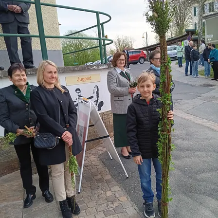 Eine Gruppe von Erwachsenen und einem Kind hält eine große Pflanze mit grünen Blättern auf einer Stadtstraße. Es sind Autos am Straßenrand geparkt und Menschen gehen vorbei. Ein Schild hinter ihnen lautet 'Jugend'.