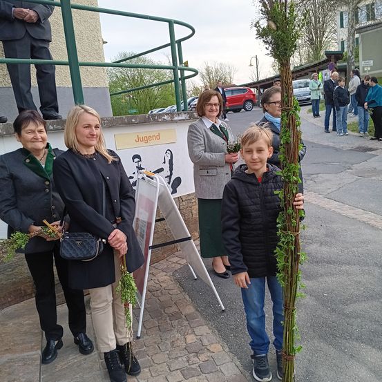 Eine Gruppe von Erwachsenen und einem Kind hält eine große Pflanze mit grünen Blättern auf einer Stadtstraße. Es sind Autos am Straßenrand geparkt und Menschen gehen vorbei. Ein Schild hinter ihnen lautet 'Jugend'.