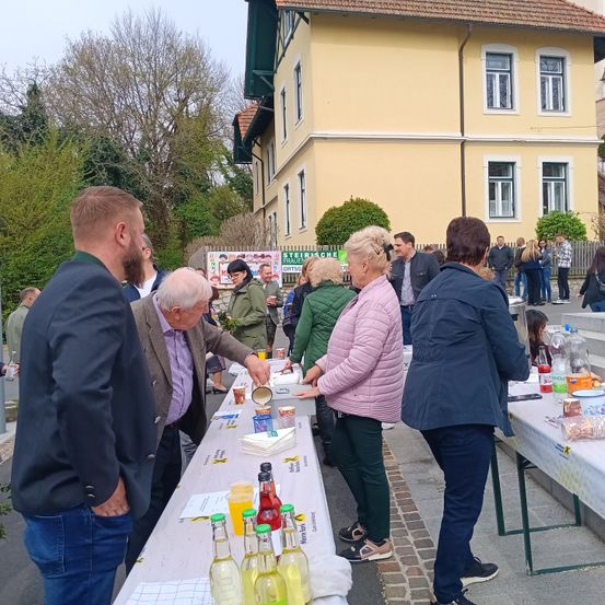 Eine Gruppe von Menschen steht vor einem langen Tisch im Freien vor einem gelben Gebäude. Der Tisch ist mit einem weißen Tuch bedeckt und hat Flaschen, Tassen und andere Gegenstände darauf. Ein Mann gießt etwas in einen Becher. Hinter ihnen stehen Menschen in der Nähe eines Zauns.