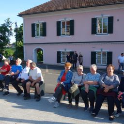 Eine Gruppe älterer Frauen und Männer sitzt auf einer Bank vor einem rosa Gebäude mit grünen Fensterläden.