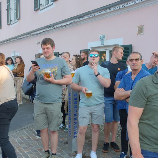 Mehrere Menschen stehen in einer Schlange und halten Gläser mit Bier. Sie stehen vor einem Gebäude mit einem Schild, auf dem 'Trinkbrunnen' steht. Der Mann auf der linken Seite hält ein Handy und schaut darauf, während der Mann neben ihm eine Zigarette in der Hand hat. Eine Frau steht links, und einige Leute stehen hinter ihnen.