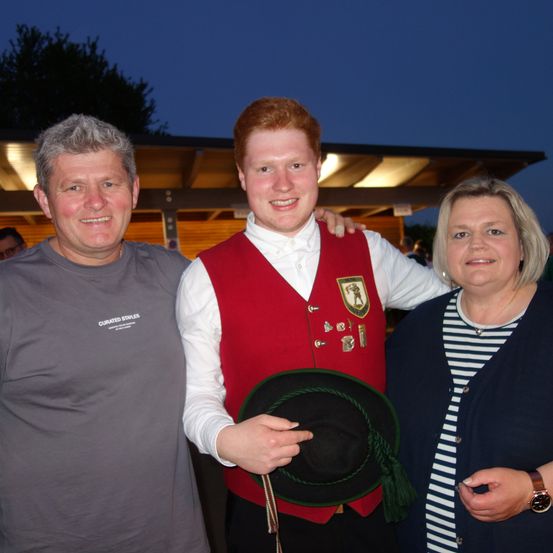 Drei Menschen stehen bei Nacht draußen. Der Mann links trägt ein graues T-Shirt mit weißem Text. Der Mann in der Mitte trägt ein rotes Weste und hält einen schwarzen Hut. Die Frau rechts trägt eine blaue Strickjacke und eine Halskette. Ein hölzernes Gebäude mit Lichtern ist im Hintergrund.
