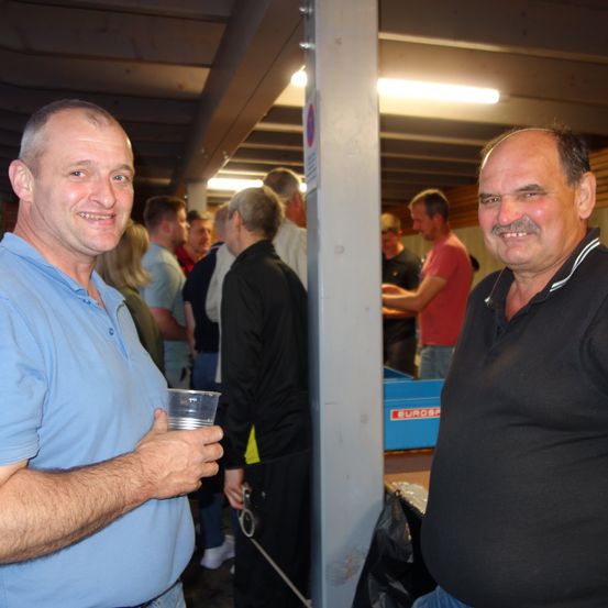 Zwei Männer lächeln und halten einen Becher bei einer Indoor-Versammlung. Hinter ihnen stehen einige Leute und unterhalten sich. Ein blauer Kasten steht auf dem Boden, und einige Lampen sind an der Decke montiert.