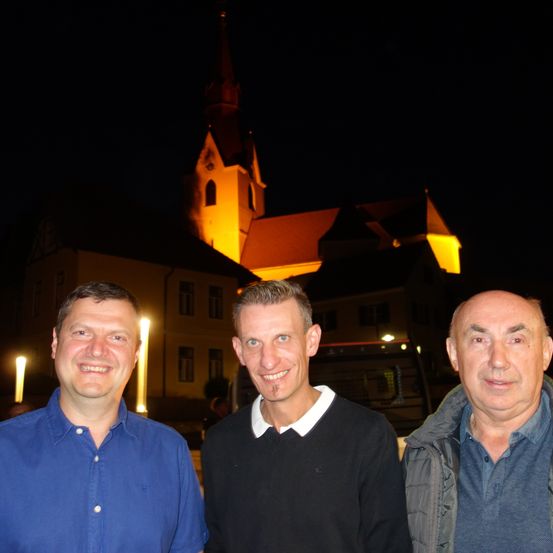 Drei Männer lächeln in die Kamera vor einer Kirche bei Nacht. Die Kirche ist beleuchtet.