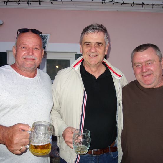 Drei Männer stehen eng beieinander und lächeln. Der Mann auf der linken Seite hält einen großen Becher mit Bier, während der Mann in der Mitte ein kleineres Glas hält.