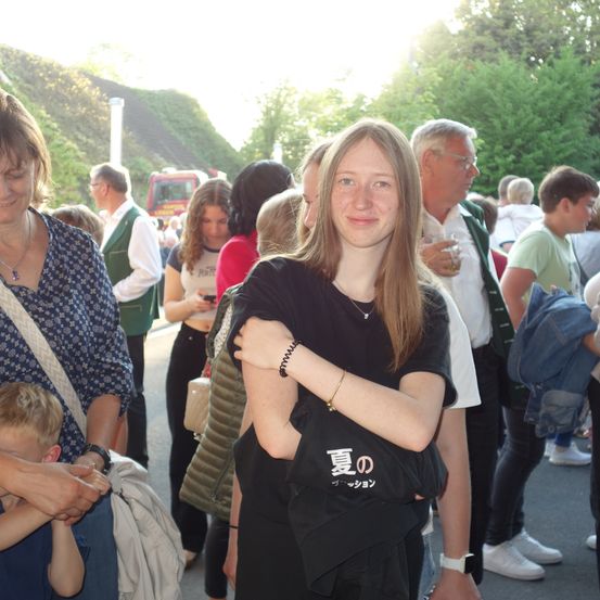 Eine Frau mit langen blonden Haaren, die ein schwarzes T-Shirt trägt und eine schwarze Tasche hält, lächelt inmitten einer Menschenmenge.