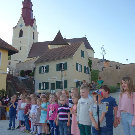 Eine Gruppe von Kindern steht vor einem Kirchengebäude mit Turm. Sie singen wahrscheinlich.