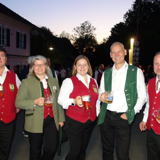 Fünf Personen posieren mit Biergläsern lächelnd für ein Foto. Sie tragen traditionelle Kleidung. Hinter ihnen stehen einige Personen, und es gibt Bäume und Gebäude im Hintergrund.