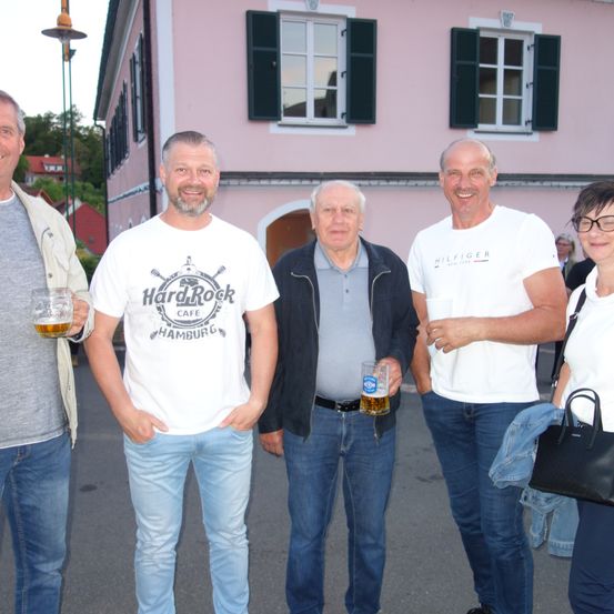 Fünf Männer stehen lächelnd vor einem rosa Gebäude, einer hält ein Glas Bier. Das Gebäude hat drei Fenster und eine schwarze Tür.