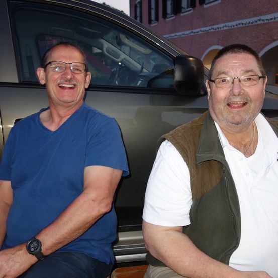 Zwei Männer sitzen vor einem Auto und lächeln in die Kamera. Einer trägt ein blaues Shirt, der andere ein weißes Shirt mit einer braunen Weste.