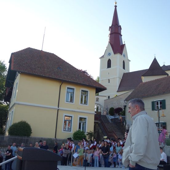 Ein Mann spricht in ein Mikrofon vor einer Menschenmenge, die sich vor einer Kirche mit einem Kirchturm und einer Uhr versammelt hat.