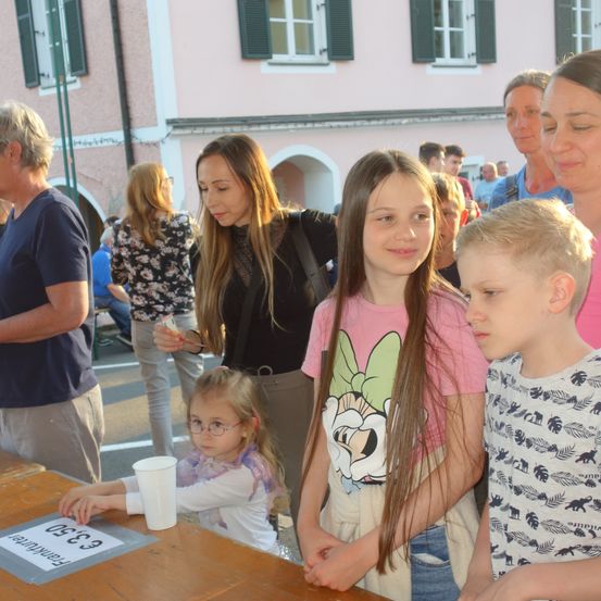 Eine Gruppe von Menschen steht bei einem Tisch bei einer Veranstaltung im Freien. Ein Mädchen hält einen Becher, während ein anderes Mädchen in die Kamera lächelt.