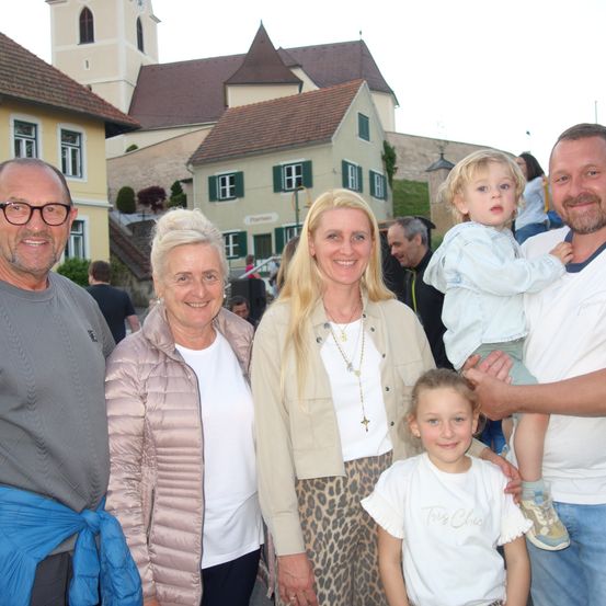 Eine Familie mit vier Erwachsenen und zwei kleinen Kindern posiert für ein Foto vor einem europäischen Dorf. Die Erwachsenen lächeln und einer hält ein Kind auf seinen Schultern. Das Kind lächelt ebenfalls.