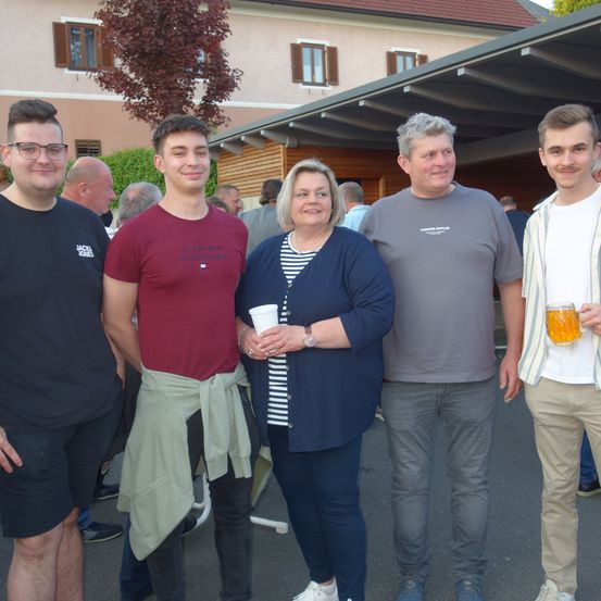 Fünf Menschen stehen an einem sonnigen Tag draußen, mit Gebäuden im Hintergrund. Die Frau in der Mitte hält einen Becher, und der Mann rechts hält ein Glas.