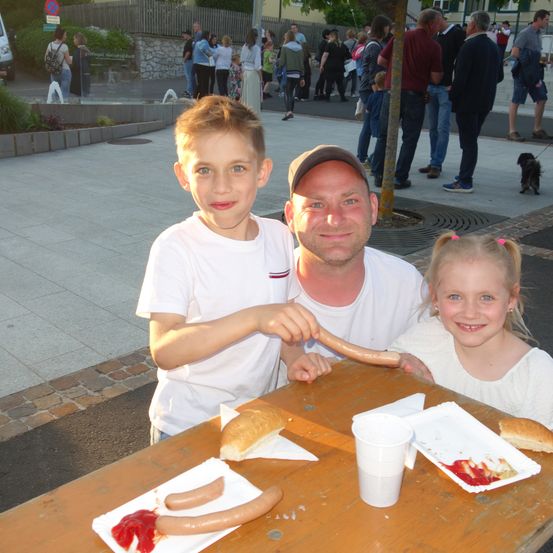 Ein Vater und seine zwei Kinder sitzen draußen an einem Tisch und genießen eine Mahlzeit. Auf dem Tisch befinden sich eine Hotdog, ein Brötchen und Ketchup. Hinter ihnen gehen Menschen auf dem Bürgersteig, und ein Baumstamm ist in der Nähe.