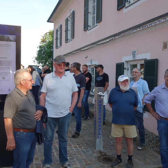 Gruppe von Männern steht draußen vor einem rosa Gebäude mit einem Wasserbrunnen, der Trinkbrunnen heißt.