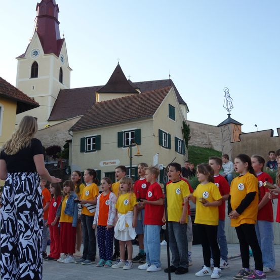Eine Gruppe von Kindern in passenden gelben und roten Shirts steht vor einem Kirchengebäude und singt, während eine Frau in Schwarz hinter ihnen steht. Die Kirche hat einen Glockenturm, und es gibt eine Statue einer Frau auf dem Dach der Kirche. Daneben stehen auch andere Menschen.