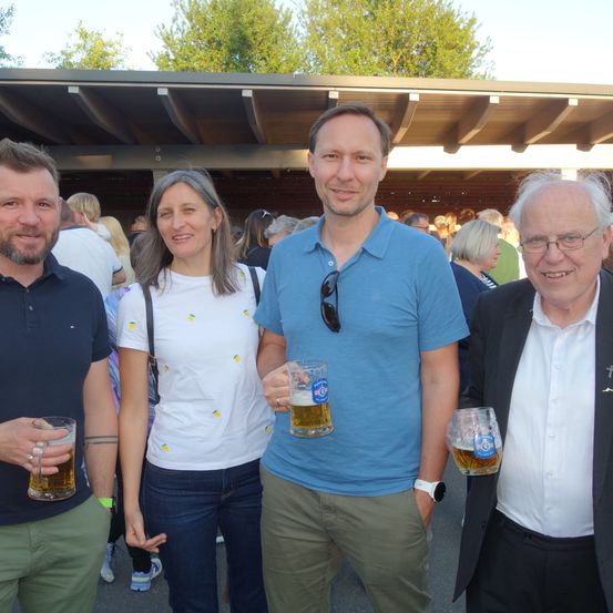 Vier Personen stehen nebeneinander und halten ein Bierglas. Drei von ihnen lächeln. Der Mann rechts trägt eine Brille.