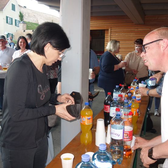 Eine Frau in einem schwarzen Shirt bereitet sich darauf vor, an einem Tisch mit verschiedenen Getränkeflaschen und Bechern zu bezahlen, während ein Mann in einem weißen Shirt eine Plastikflasche hält.