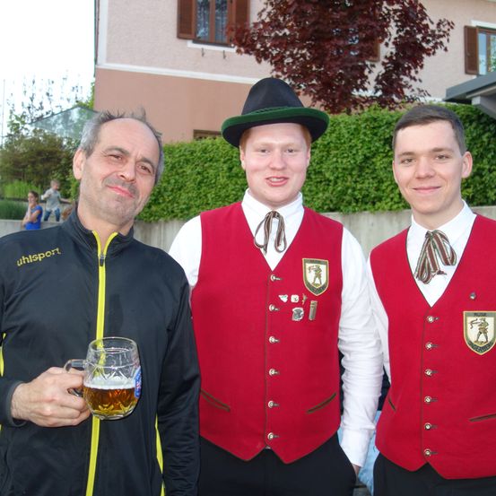 Drei Männer stehen vor einem Gebäude. Zwei Männer tragen rote Westen, und der Mann auf der linken Seite trägt eine schwarze Jacke und hält ein Glas Bier.