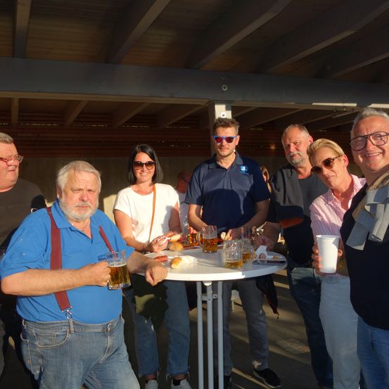 Eine Gruppe von Menschen versammelt sich um einen Tisch mit Biergläsern und Essen. Sie lächeln und posieren für ein Foto.