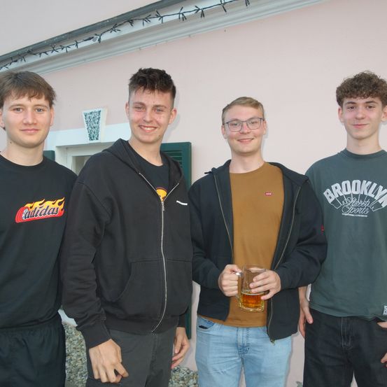 Vier junge Männer stehen vor einem rosa Gebäude. Einer hält ein Glas Bier, während die anderen Kapuzenpullover und T-Shirts tragen.