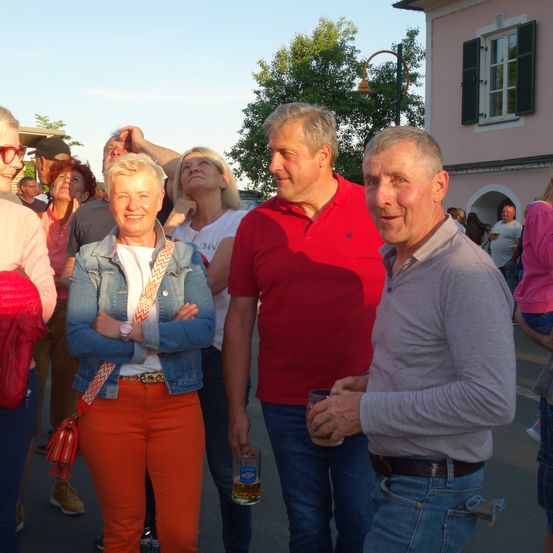 Gruppe von Menschen, die draußen in der Nähe eines Gebäudes stehen und reden. Eine Frau mit orangefarbener Hose und einer Jeansjacke lächelt. Ein Mann mit rotem Hemd und einem Glas Bier steht neben ihr.
