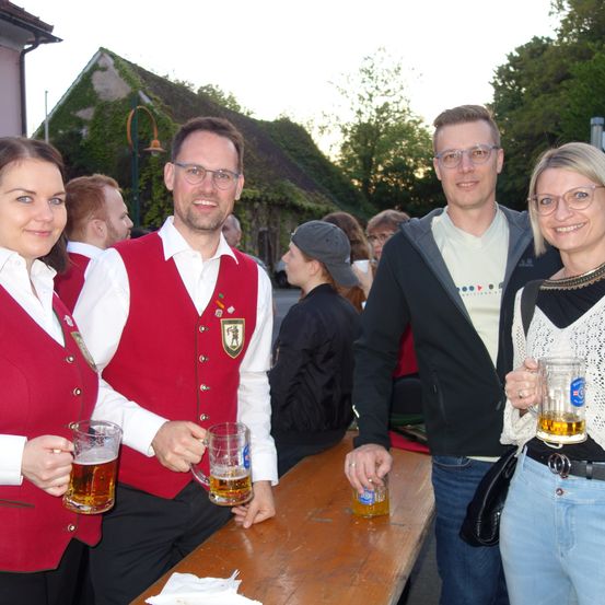 Vier Erwachsene, zwei Männer und zwei Frauen, stehen hinter einem Holztisch mit Bierkrügen in den Händen. Sie lächeln und posieren für ein Foto.