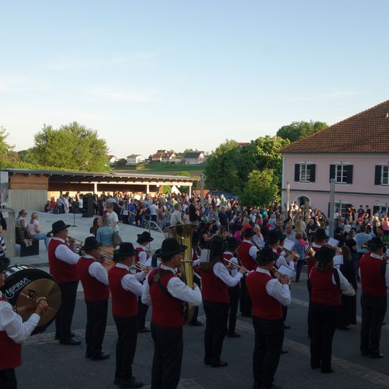 Ein Blasorchester spielt vor einer Menschenmenge bei einem Tagesevent. Die Band trägt rote Westen und schwarze Mützen. Das Publikum sitzt auf dem Bürgersteig.