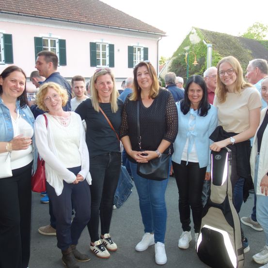 Eine Gruppe lächelnder Frauen und Männer steht auf einer Straße, mit einem rosa Gebäude und grünen Fensterläden dahinter. Sie nehmen wahrscheinlich an einer Veranstaltung teil, da eine Frau eine Gitarrenkoffer hält.
