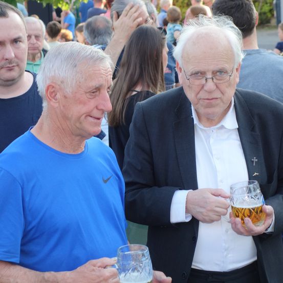 Zwei ältere Männer, einer in einem blauen Shirt und der andere in einem dunklen Anzug, halten Biergläser und lächeln in einer überfüllten Außenumgebung.