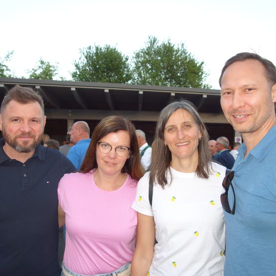 Vier Personen lächeln und posieren für ein Foto vor einem Gebäude mit Bäumen im Hintergrund.