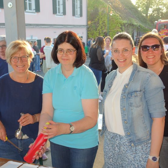 Vier Frauen lächeln und posieren für ein Foto. Die Frau auf der linken Seite hält ein rotes Objekt, möglicherweise eine Flasche. Die Frau neben ihr hält eine Flasche und einen Löffel. Sie stehen vor einem Gebäude, in dem viele Menschen herumlaufen.