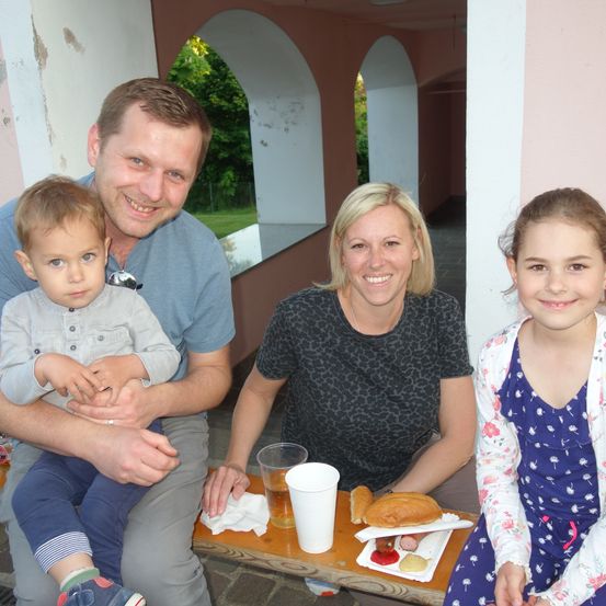 Eine Familie sitzt auf einer Bank im Freien, lächelt und posiert für ein Foto. Der Mann hält ein Baby und eine Frau sitzt neben ihm. Ein junges Mädchen sitzt rechts und ein Holztisch vor ihnen hat Essen und Getränke.