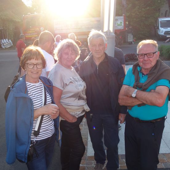 Vier Menschen stehen draußen und lächeln, einer der Männer trägt eine Brille und einen Schal. Hinter ihnen ist ein helles Licht, und im Hintergrund sind Bäume, ein Gebäude und ein Feuerwehrwagen zu sehen.