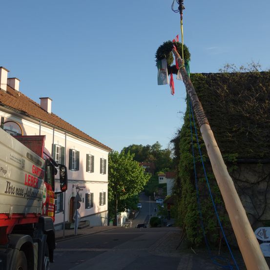 Ein Lkw ist am Straßenrand geparkt, mit einem mit Flaggen geschmückten Holzpfahl im Vordergrund. Der Pfahl ist mit einem Seil an einem Lkw befestigt. Ein Gebäude mit einem braunen Dach steht hinter dem Lkw, mit einigen Fenstern und einer Lampe. Bäume und Pflanzen befinden sich am Straßenrand.