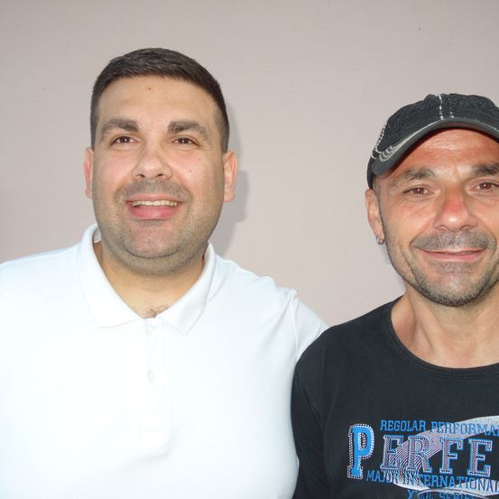 Zwei Männer stehen eng beieinander, einer trägt ein weißes Poloshirt und der andere ein schwarzes T-Shirt mit blauem Text, der 'REGULAR PERFORMANCE' und 'PERFE' lautet. Sie lächeln beide und schauen in die Kamera.